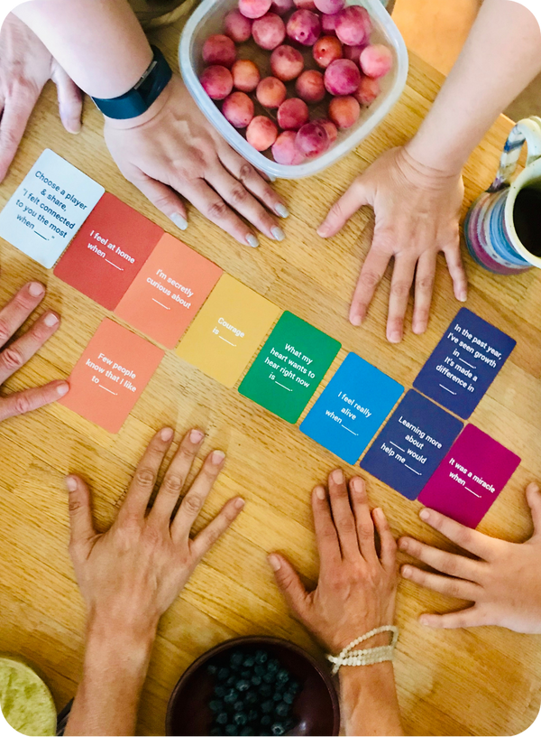 Hands reaching towards Cards-for-Connection cards on a wooden table with a bowl of fruit in the background, in a home setting.