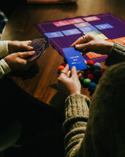 Load image into Gallery viewer, Hands holding Cards for Connection during gameplay at a wooden table on a purple mat.