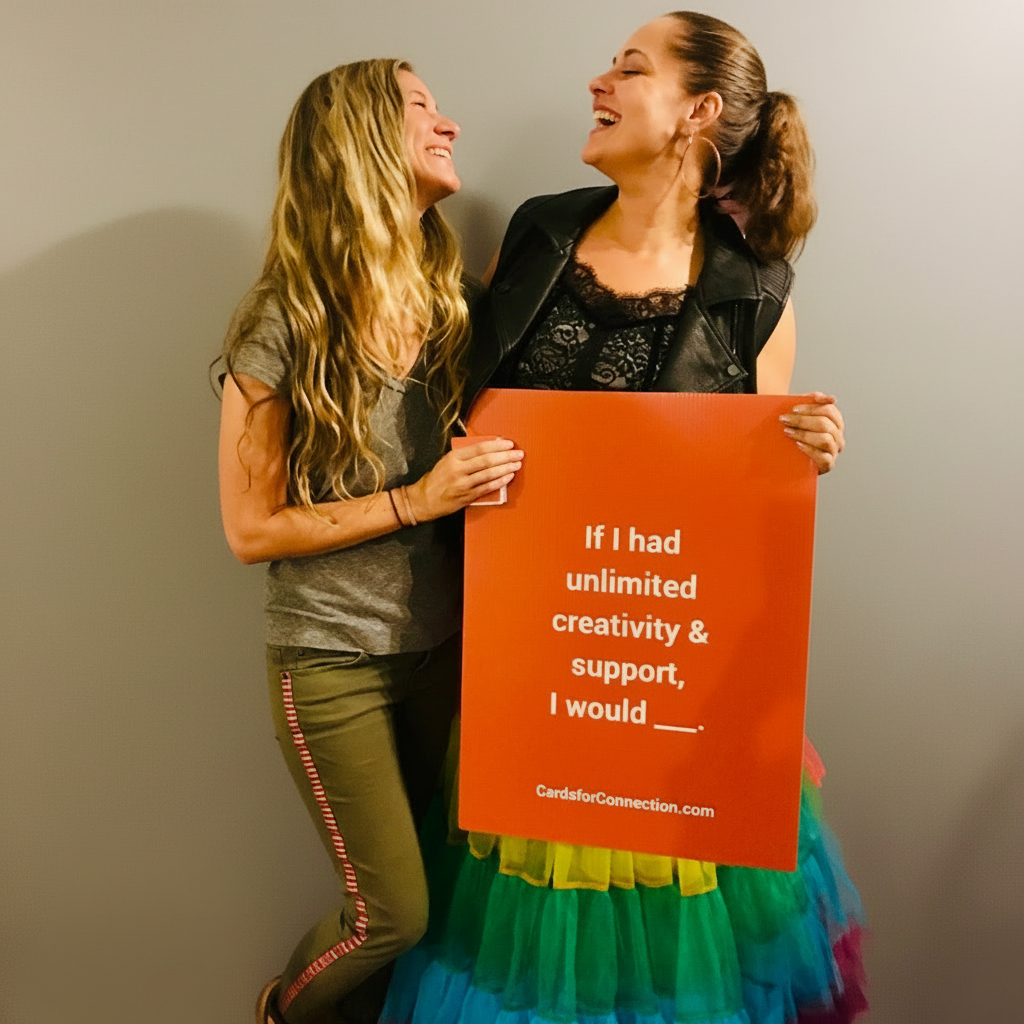 Two women standing together, one holding an orange sign that reads "if i had unlimited creativity and support, I would ___"